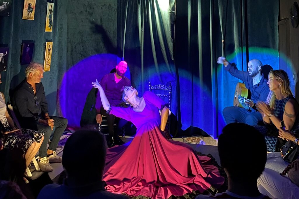 Bailarina en un escenario íntimo, vistiendo un vestido morado, realizando una pose elegante mientras el guitarrista toca en el fondo, con luces suaves iluminando la escena