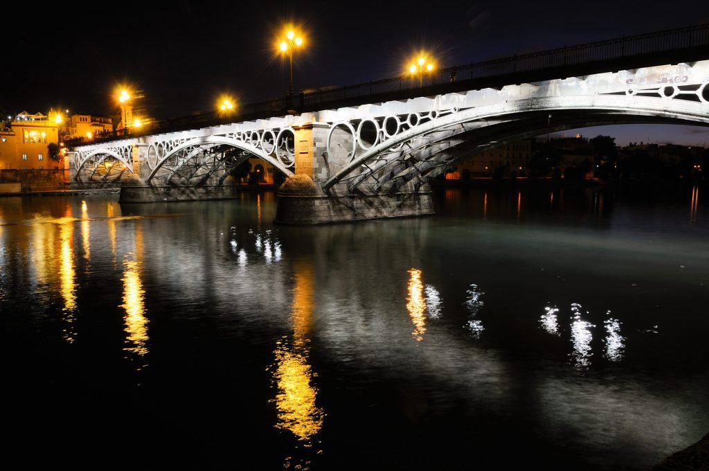El Puente de Triana en Sevilla iluminado por la noche, reflejado sobre el río Guadalquivir.