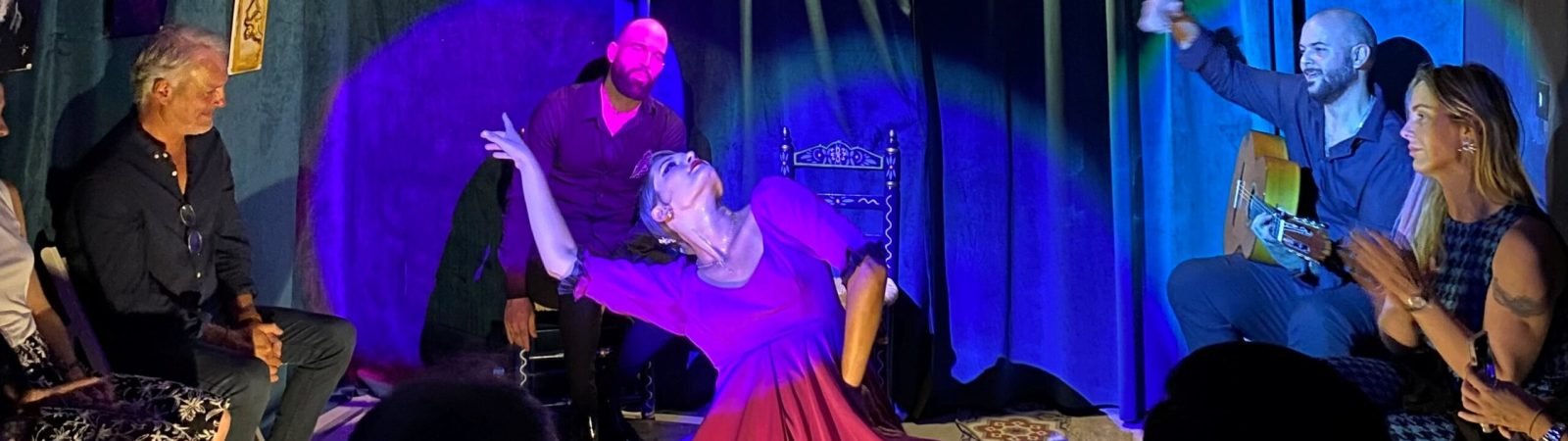 Bailarina en un escenario íntimo, vistiendo un vestido morado, realizando una pose elegante mientras el guitarrista toca en el fondo, con luces suaves iluminando la escena