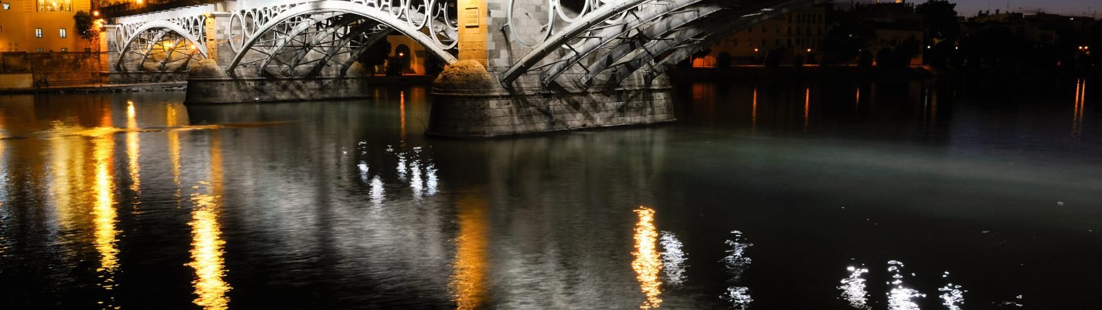 El Puente de Triana en Sevilla iluminado por la noche, reflejado sobre el río Guadalquivir.